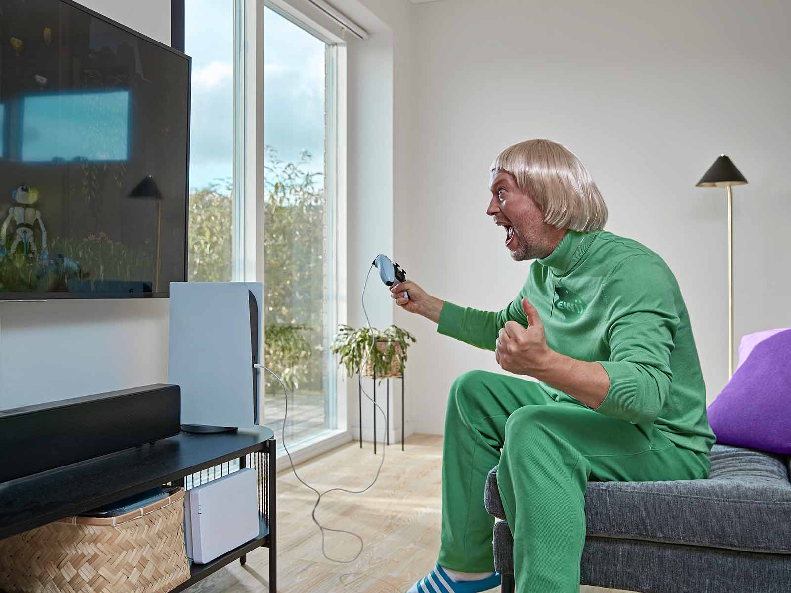 Mand i grønt tøj sidder på sofa med gaming controller og jubler over at han har bestilt 1000 Mbit fibernet til 0 kr. hos EWII.