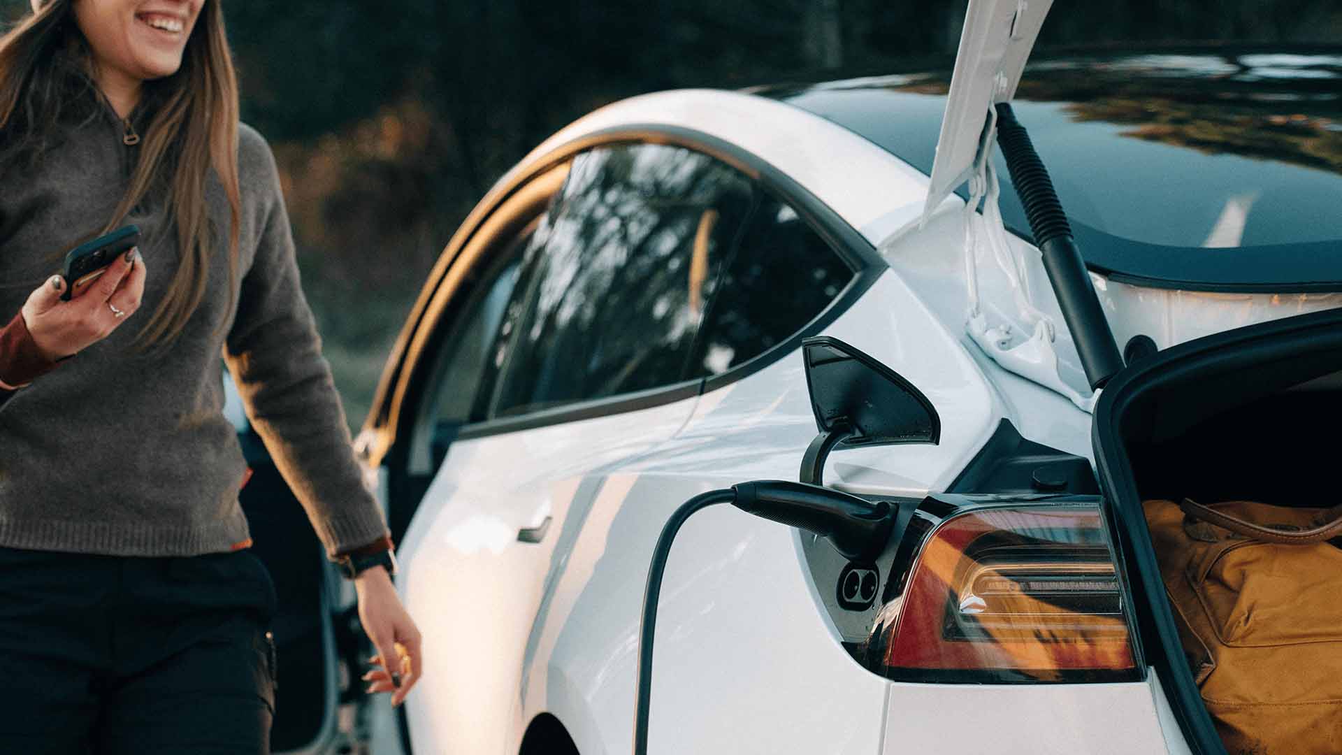 Kvinde med mobil ved elbil med ladekabel. Hun er ved at undersøge hvad koster det at lade en elbil. Svaret afhænger bl.a. om du lader ude eller hjemme. Se mere her.